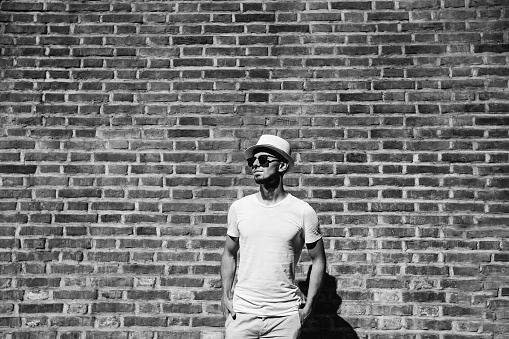 guy with beard in white t-shirt and sunglasses hold hands in pockets. the guy stands near the brick wall. black and white photo