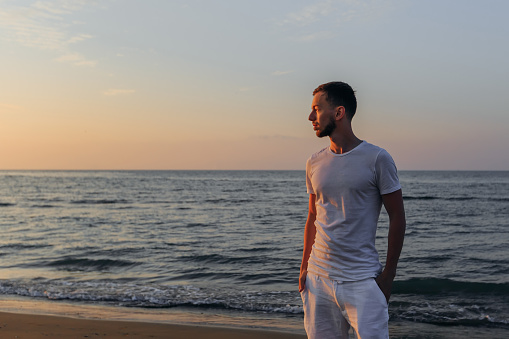 a guy in a white T-shirt and shorts holds his hands in his pockets on the sandy beach by the sea