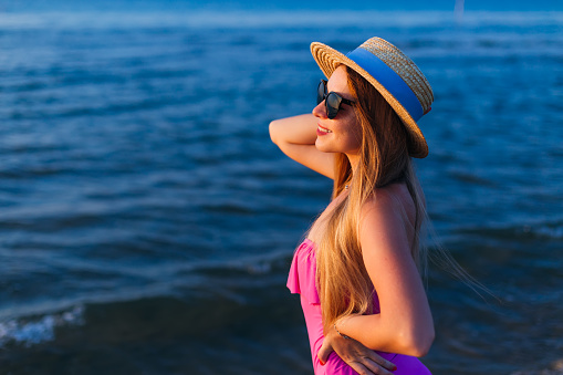 young woman in boater and pink swimming costume has charming smile on face. looking at the sea
