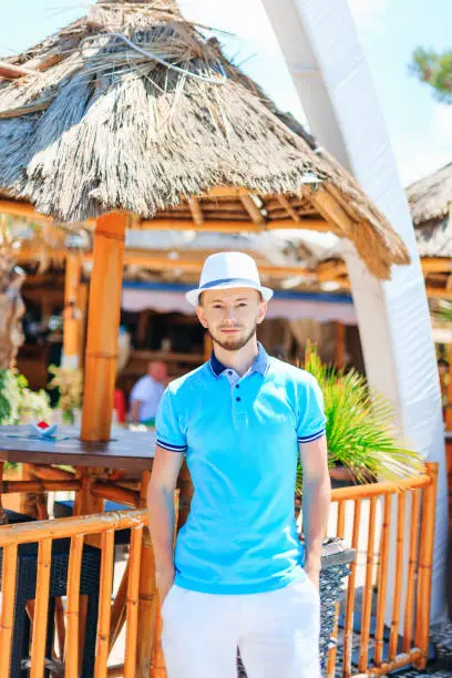 Photo of A young man wearing a boater and polo standing near cafe. The gu