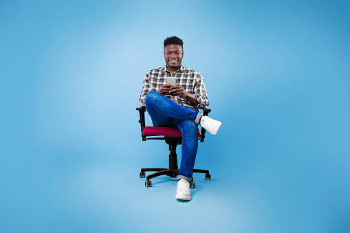 Handsome young black guy using cellphone, surfing web or social media, sitting on chair, enjoying contemporary technologies, checking new mobile app on blue studio background