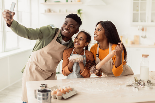 Cheerful African American Family Baking Cookies And Making Selfie On Mobile Phone Holding Dough And Embracing Standing In Modern Kitchen At Home On Weekend. Pastry Recipes Concept