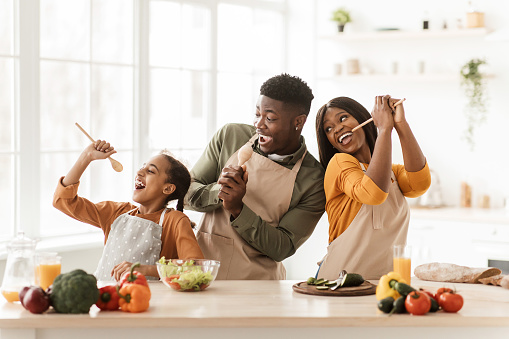 Positive Black Family Of Three Cooking And Having Fun Singing Favorite Song Holding Spoons Preparing Dinner Together Standing In Modern Kitchen Indoor. Weekend Leisure, Healthy Food Recipes Concept