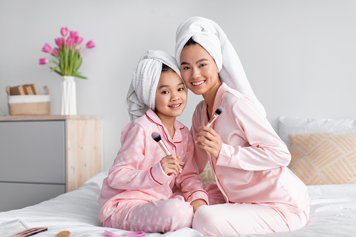 Smiling young pretty japanese female and teenage girl in pajamas and towel sit on bed with makeup brushes in bedroom interior. Mom and daughter and beauty care at home, during covid-19 quarantine