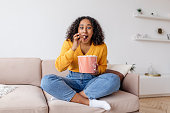 Excited young African American woman sitting on couch, watching TV and eating popcorn at home
