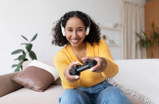 Happy millennial black woman in headphones using joystick to play videogame on sofa at home. Joyful African American lady in headset having fun with computer game. Domestic hobbies