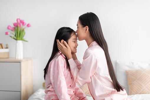 Cheerful japanese millennial female kissing at forehead of teenage girl in bedroom interior, profile, free space. Family relationship, mom and daughter enjoy tender moment together, relationships