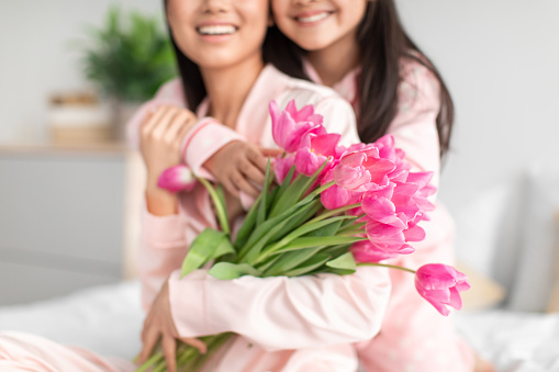 Glad teenage girl hugging millennial japanese lady in pajama giving bouquet of tulips celebrating mother's day in bedroom. Fun together, daughter congratulates mom with birthday at home due covid-19