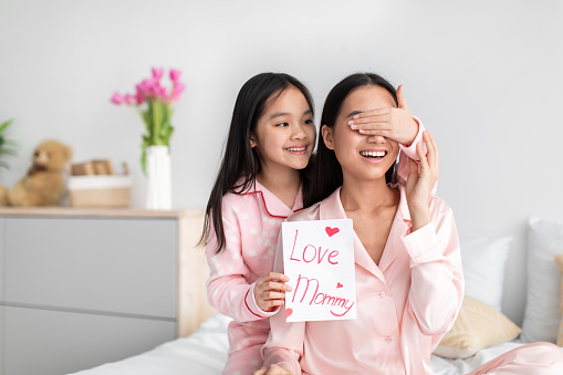 Smiling asian cute teen girl in pajama closes eyes to millennial woman hold postcard in bedroom interior, daughter congratulate mom. Surprise at mothers day, celebration at home due covid-19 outbreak