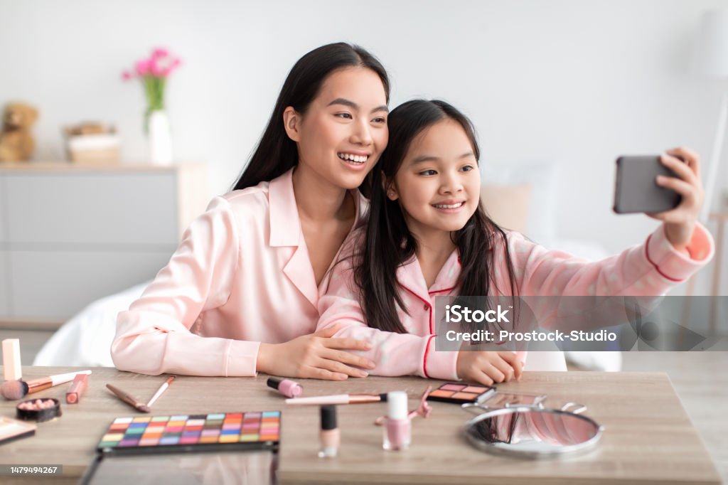 Smiling japanese teen daughter and young mom in pajamas at table take selfie for beauty blog in bedroom Smiling japanese teen daughter and young mom in pajamas at table take selfie for beauty blog in bedroom interior. App, device, social networks. Love and relationships, skin care and photo at home 10-11 Years Stock Photo