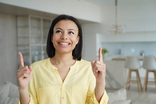 Happy woman look up pointing up with fingers. Smiling excited female client customer showing special online store offer, holding finger up, demonstrating new product, amazing news, standing at home.