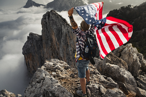 Proud man raising the flag of the United States of America standing on the top of the mountain above beautiful clouds. Successful climb to the top of the mountain.