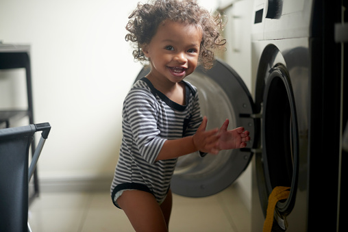 Happy, smile and baby playing by a washing machine for adventure, freedom and fun at a house. Happiness, naughty and portrait of a excited boy toddler child by a washer in the laundry room at home.