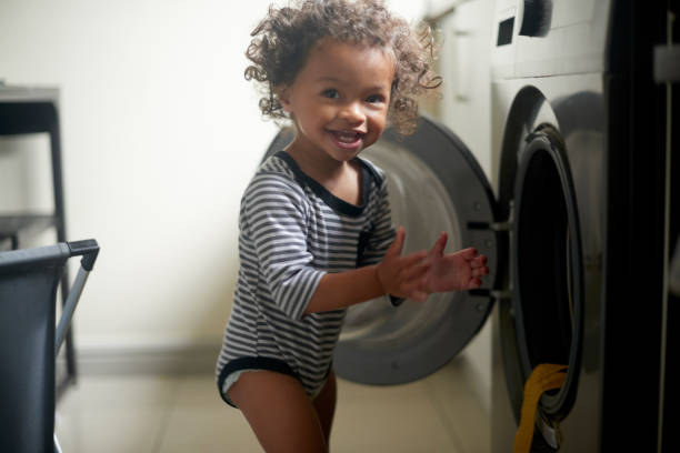 Happy, smile and baby playing by a washing machine for adventure, freedom and fun at a house. Happiness, naughty and portrait of a excited boy toddler child by a washer in the laundry room at home. stock photo