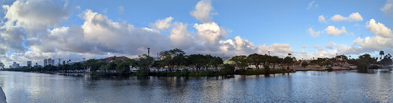 The Ala Wai Canal is a man-made waterway that runs through the city of Honolulu on the island of Oahu. It is lined with condos, golf courses, and coconut trees. In the distance, the mountains of the Ko'olau Range can be seen. This panoramic photo shows the beauty and tranquility of this popular tourist destination.