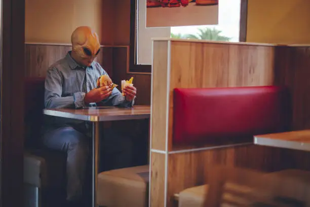 Photo of One man with alien mask eating alone inside a fast food store bad food hamburger and french fries. Ufo are among us living as human concept funny image. Invasion and diversity people indoor leisure