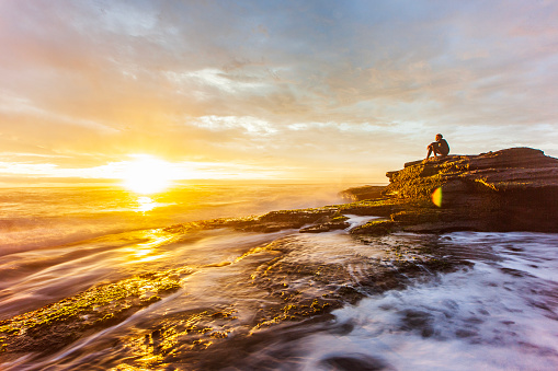Long exposure seascape of water flowing over rocks with young man observing the golden sunset. East Coast, Australia.