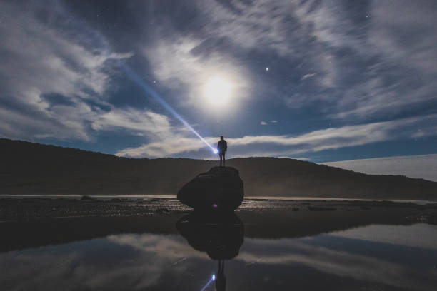 istock Man shining torch into the sky at night with a full moon with coastal landscape 1479516819