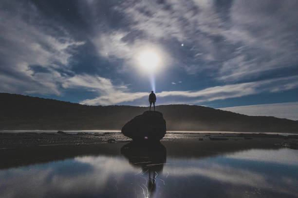 Man shining torch into the sky at night with a full moon with coastal landscape stock photo