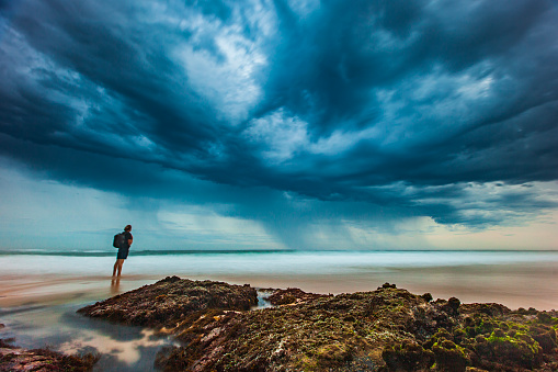 Coastal seascape scene with water flowing around rocks with man gazing at dramatic storm cloud. South East Coast, Australia.