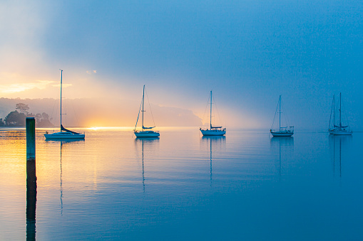 Sailing boats in calm bay with thick fog and morning mist at sunrise. Moody atmosphere.