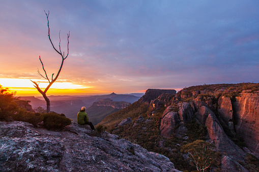 Hiker resting on rock watching sunrise over dramatic mountain landscape, South East Coast, Australia. Budawang Mountain Range.