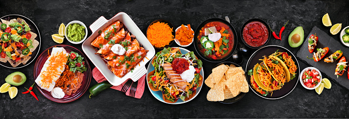 Mexican food table scene. Top down view on a dark stone banner background. Tacos, burrito plate, nachos, enchiladas, tortilla soup and salad.