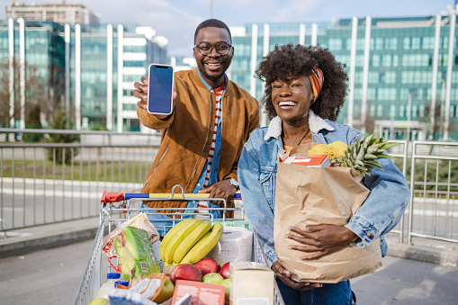 Smiling young African-American couple pulling supermarket cart and looking at the camera. Young man is holding a smart phone with blank screen