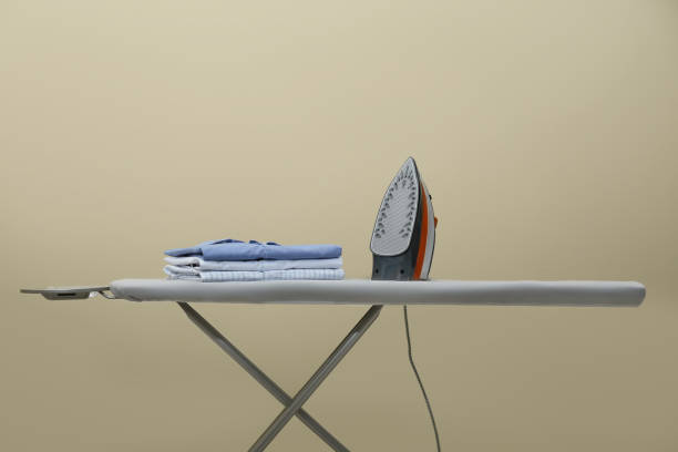 istock Modern iron and stack of clean clothes on board against beige background 1479534597