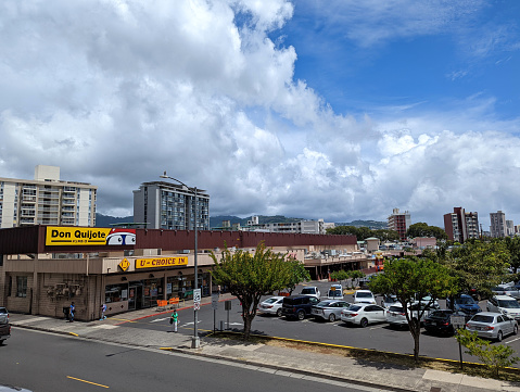 Aerial view of a Don Quijote Supermarket in Honolulu, Hawaii. The supermarket is a large, one-story building with a red and white sign. The parking lot is full of cars, and there are people walking around the area. The supermarket is located in a busy commercial area, and there are other businesses and restaurants nearby.