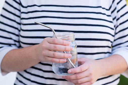 Closeup of an anonymous woman in a striped shirt holding a glass jar with a metal straw in it as a zero waste way to drink beverages by reusing the materials. She is reducing her plastic and waste impact on the environment.