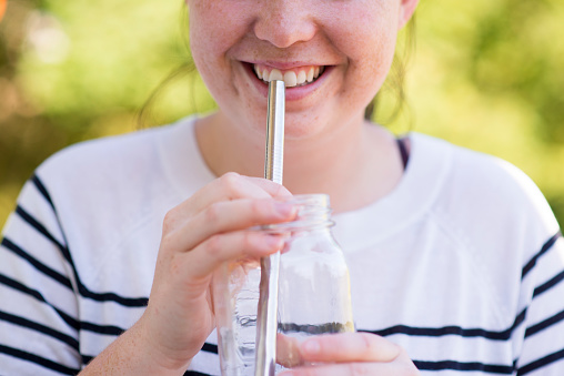 Beautiful woman in a striped white and navy shirt is smiling and holding a stainless steel metal reusable straw to her mouth as she is about to drink water from a glass jar. She is using zero waste items to reduce her impact on the environment and reduce plastic use.