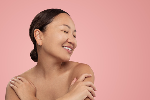 Smiling ethnic female with bare shoulders closed eyes and dark hair standing against pink background embracing herself
