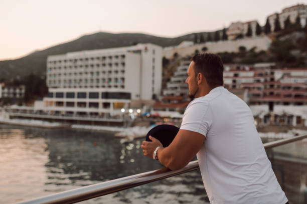 un uomo in forma e attraente in vacanza in un hotel vicino a una spiaggia sabbiosa. indossa un cappello elegante e si gode il sole caldo e il mare cristallino. lo scenario è sereno e idilliaco, ritraendo un senso di libertà, semplicità e piacere che der - clear sky water sports and fitness yacht foto e immagini stock