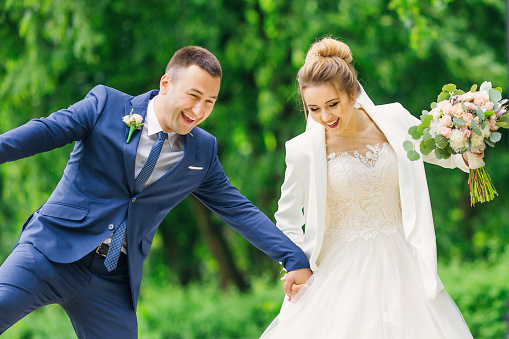 newlyweds hold hands and have fun. bride in dress holds wedding bouquet. walk in the park.