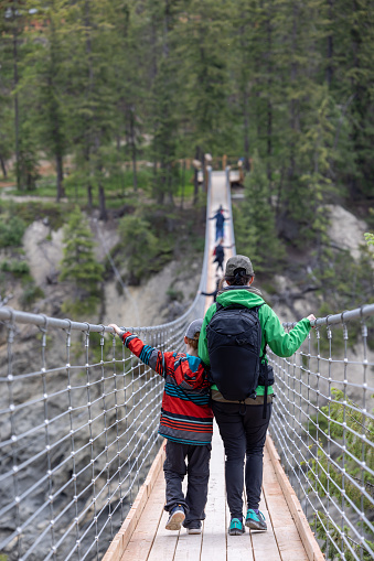 People Walking Over Wooden Suspension Bridge, British Columbia, Canada. There is a river under the bridge and The Canadian Rockies in the background.