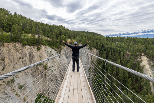 Young Man Walking Over Wooden Suspension Bridge, British Columbia, Canada. There is a river under the bridge and The Canadian Rockies in the background.