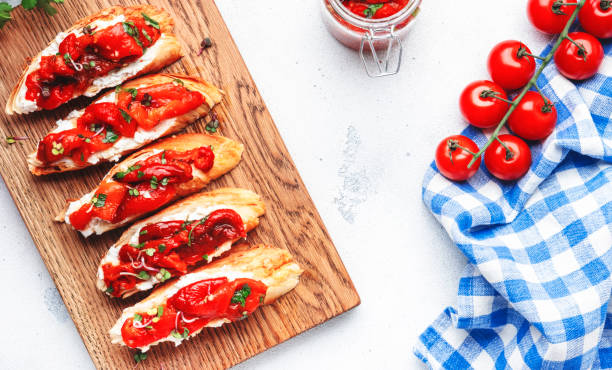Bruschetta with soft cheese and grilled red paprika in olive oil with herbs on cutting board, white kitchen table background, top view stock photo