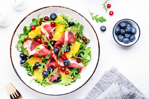 Gourmet salad with smoked duck fillet, fruit and berries, arugula and lettuce, white table background, top view