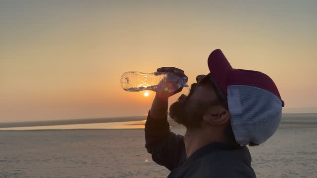 Man drinking mineral water from a white trasnparent bottle wearing sunglass and cap during sunset. Lens flair with water drinking close up on sea beach. Thirsty man taking water from bottle