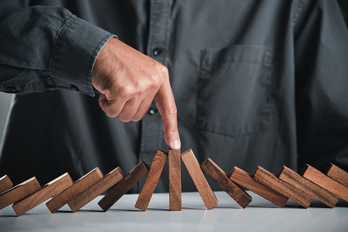 Close up hand stopping or preventing falling block in line with risk project control, Finger of businessman pointing one wooden to stopping domino effect stopped, Risk protection concept
