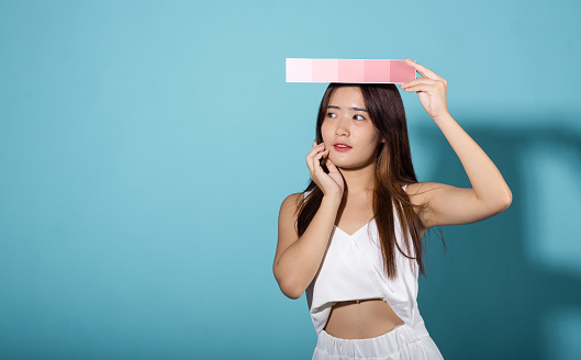 Portrait of happy female hold color card under sun protection studio shot isolated on blue background, Asian beautiful young woman smiling and holding skin color scale paper, skincare beauty face