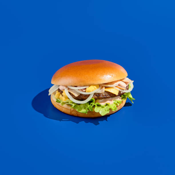 istock Tasty cheeseburger with onion, cheddar cheese, and sauce on a blue background, reflecting a modern, minimalist food photography style. Perfect for meal on-the-go and presented in a square composition 1479603488