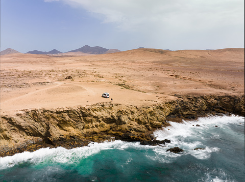 View from above, stunning aerial view a couple with a van on a cliff bathed by the ocean. Fuerteventura, Canary Islands. Life on the road concept, nomadic lifestyle.