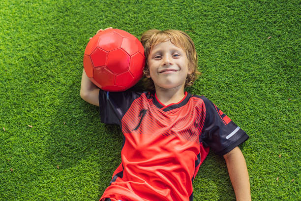 Little cute kid boy in red football uniform playing soccer, football on field, outdoors. Active child making sports with kids or father, Smiling happy boy having fun in summer stock photo