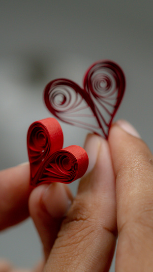 A pair of hands gently hold a quilled paper heart, the vivid red color of the heart standing out against the soft gray of the background. The sense of touch is palpable in the image, adding an intimate and personal dimension to the artwork.