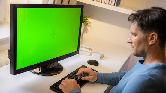 Mid adult caucasian man using computer with chroma key green screen and typing on keyboard at home