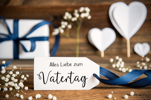 Label With German Text Alles Liebe Zum Vatertag Means Happy Fathers Day. White Festive Decoration Like Present With Blue Bow, Heart and Flowers. Vintage Wooden Background.