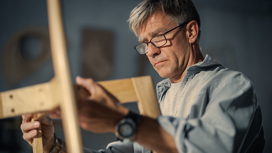 Close Up of a Carpenter Putting on Glasses, Looking at a Blueprint and Starting to Assemble a Wooden Chair. Professional Furniture Designer Working in a Studio in Loft Space with Tools on the Walls.