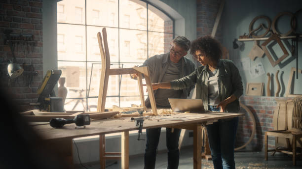 istock Small Business Owners of a Furniture Workshop Using Laptop Computer and Discussing the Design of a New Wooden Chair. Middle Age Carpenter and a Young Female Apprentice Working in Loft Studio. 1479652000